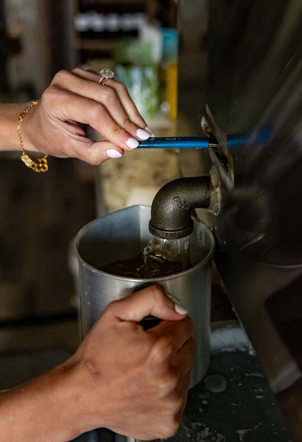 Hands with jewelry opening a metal pipe faucet valve over a steel pot to collect dark liquid.