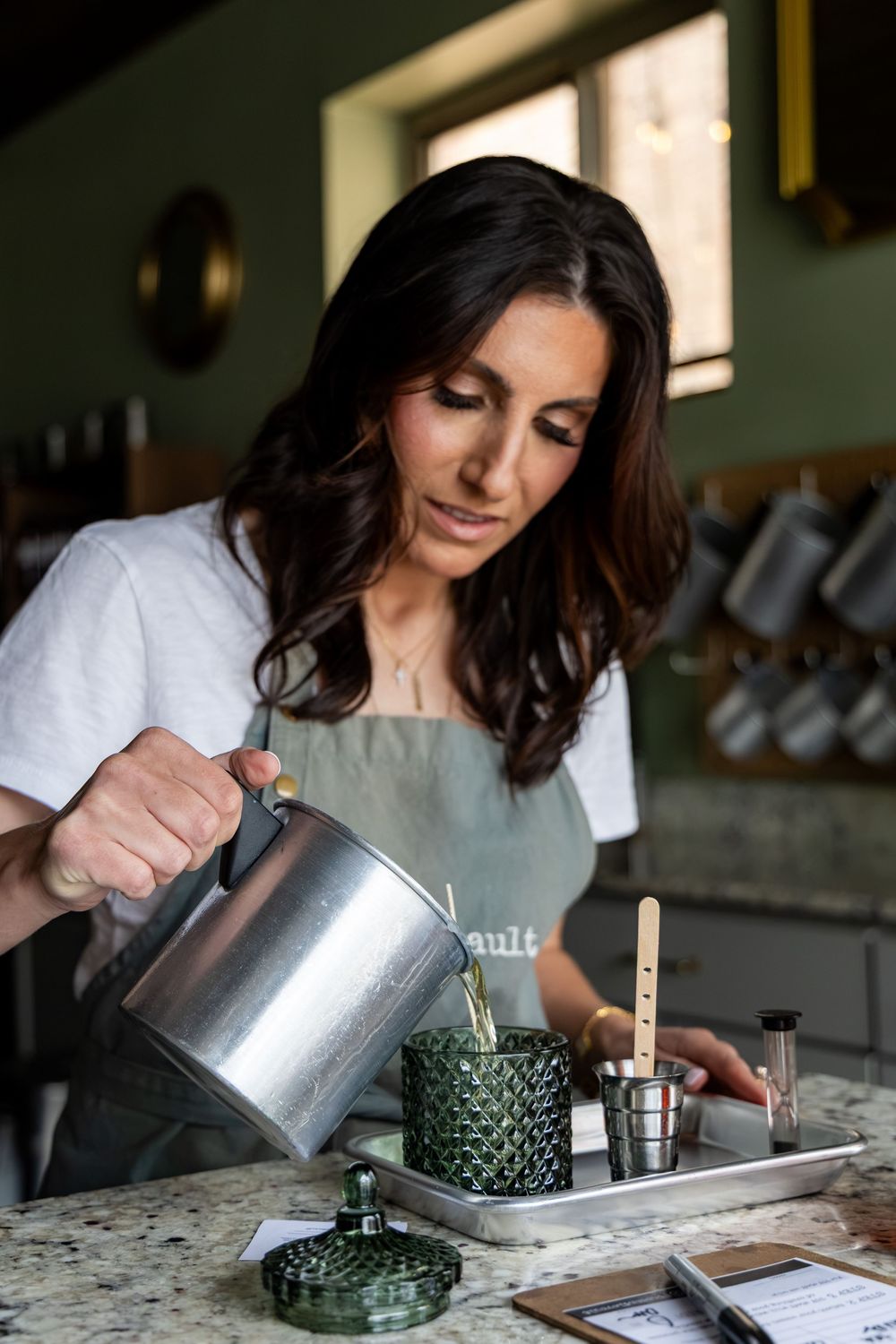 Woman in apron pouring hot water from kettle into green candle jar on marble countertop in kitchen.