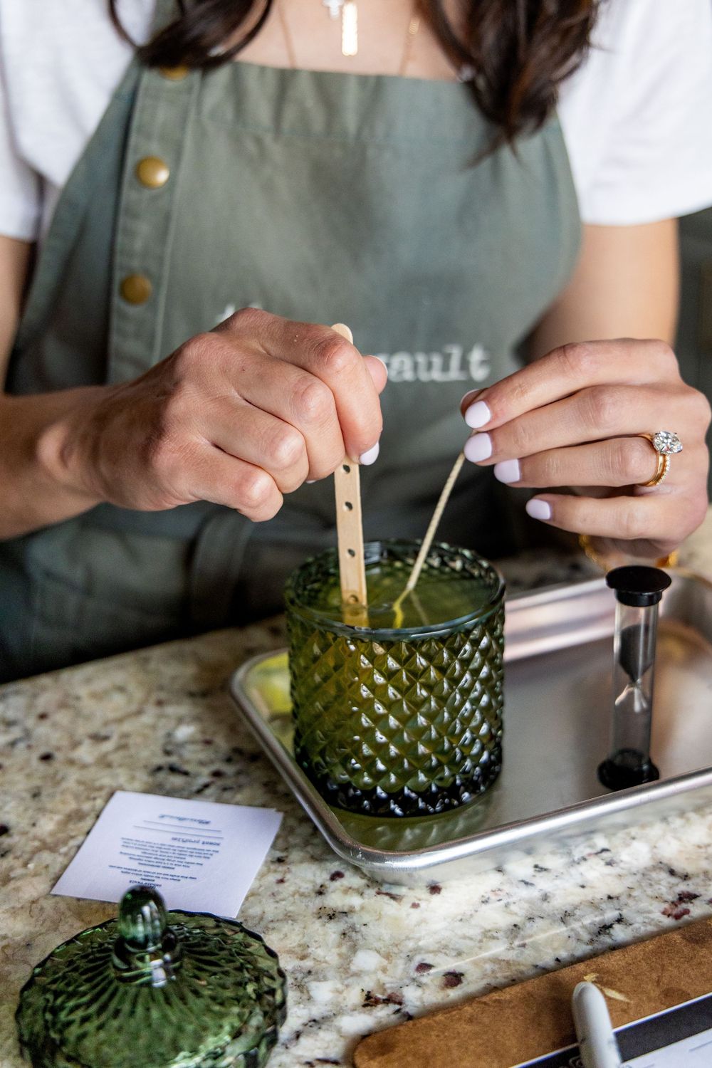 Woman in green apron making a candle, stirring wax in a green textured glass jar on a granite countertop.