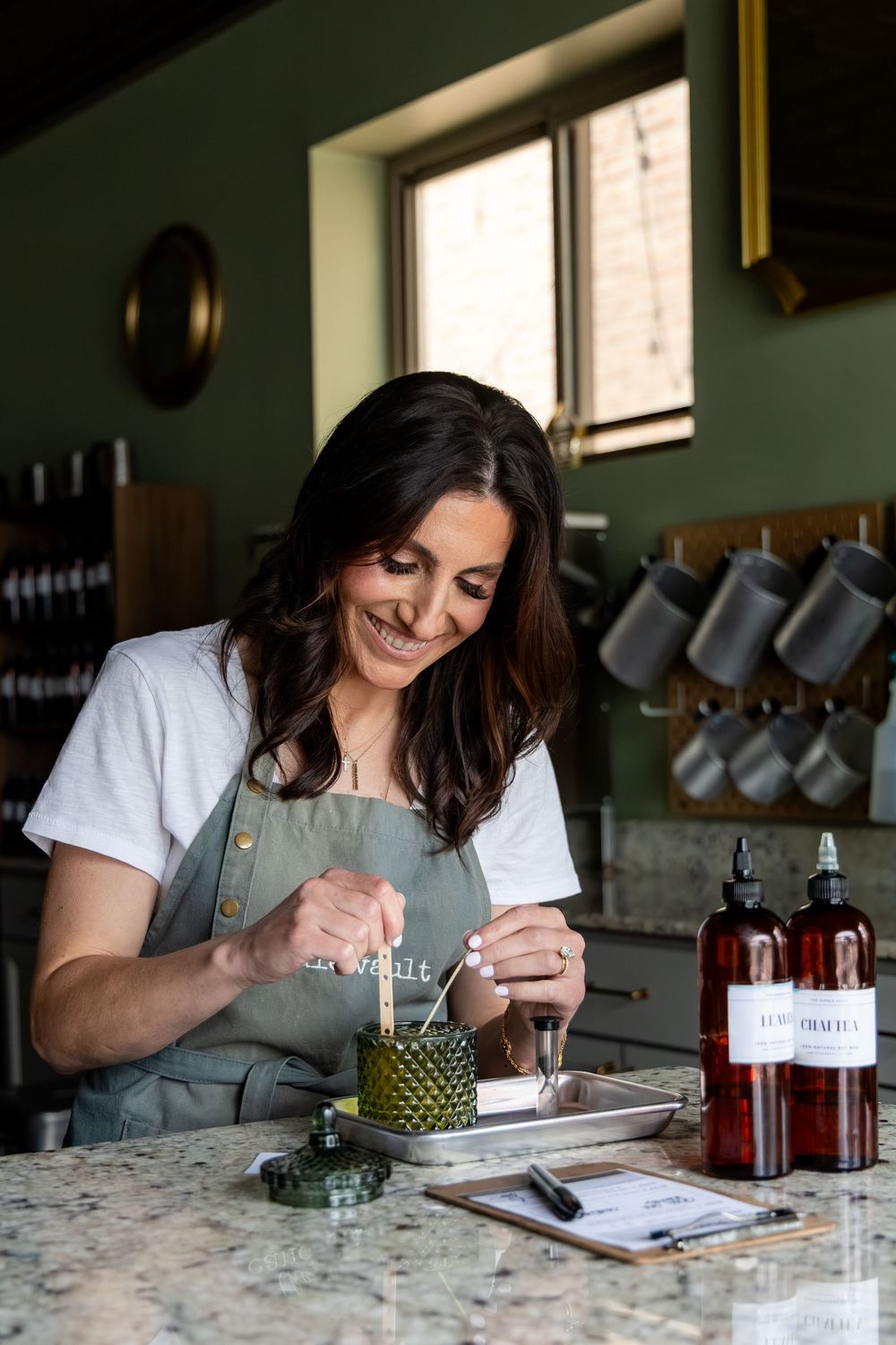 Smiling woman in green apron crafting candles at a marble counter in a stylish kitchen workshop.