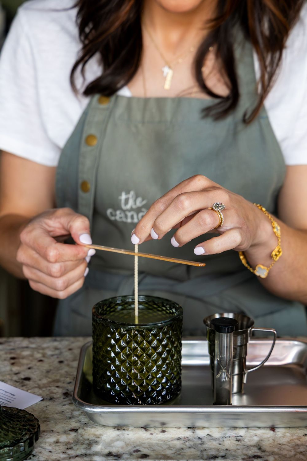 Woman in sage green apron carefully placing a wick into a dark green textured glass candle jar on a silver tray.