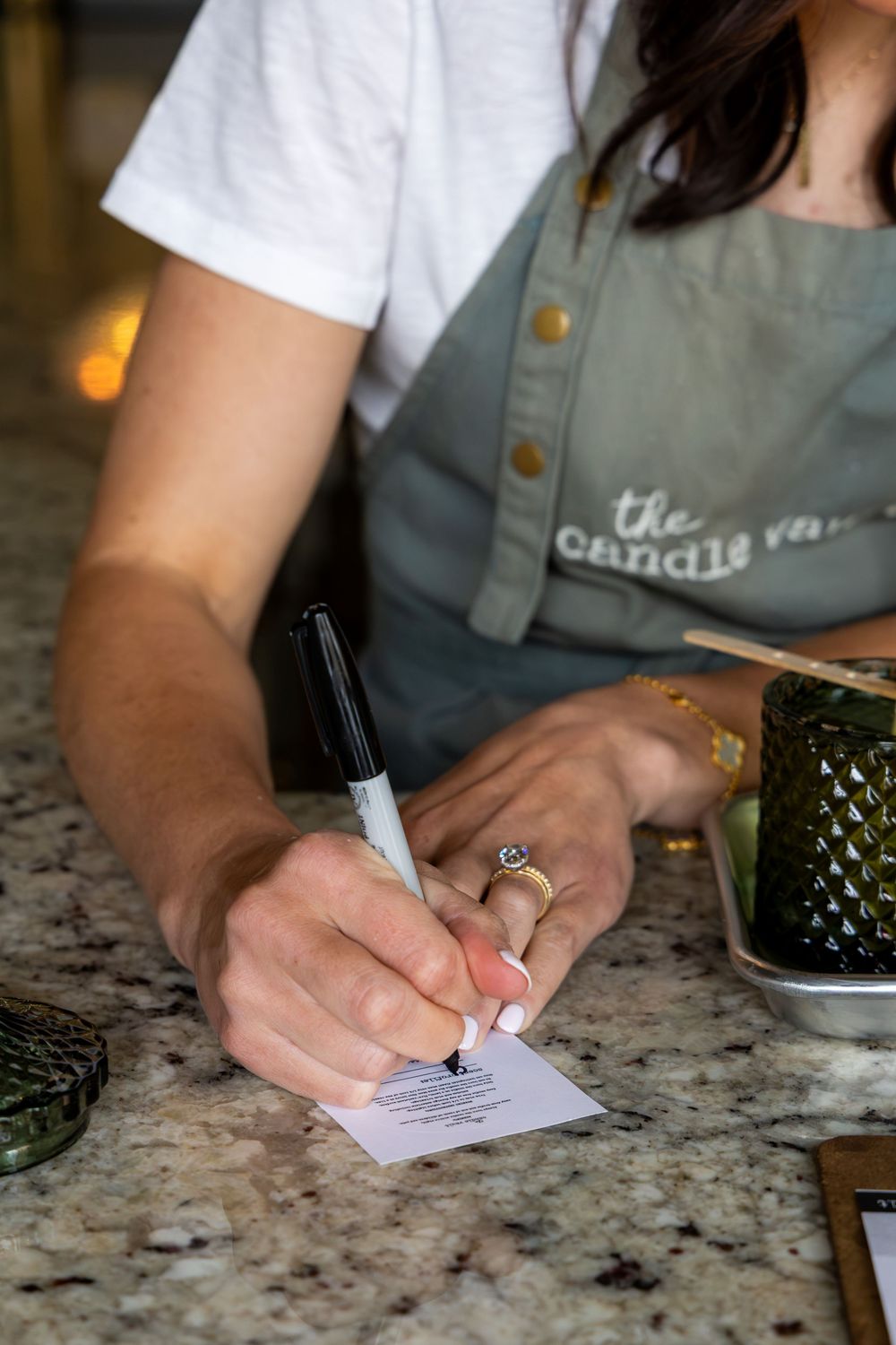 Woman in candle room apron writing on paper at granite counter with black marker.