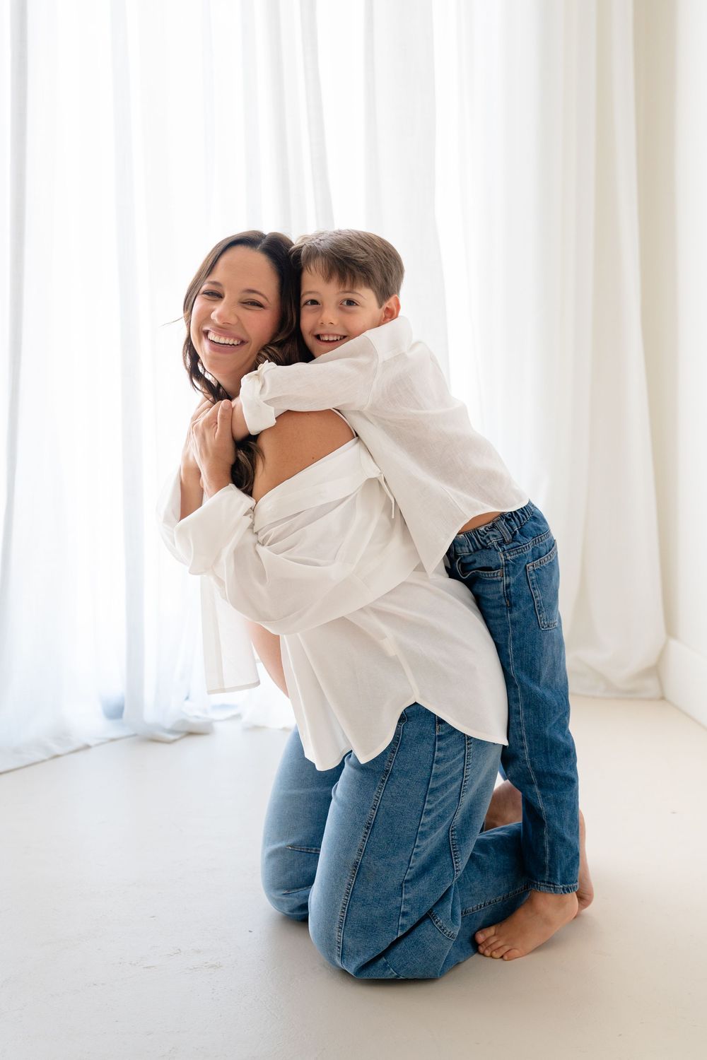 A smiling mother kneels as her young son hugs her from behind, both wearing white shirts and jeans.
