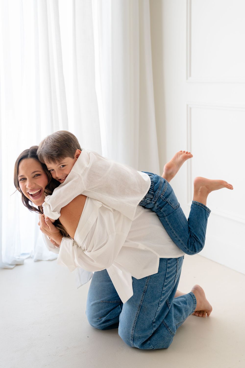 A smiling mother gives her young child a piggyback ride on the floor in a bright white room, both wearing jeans.