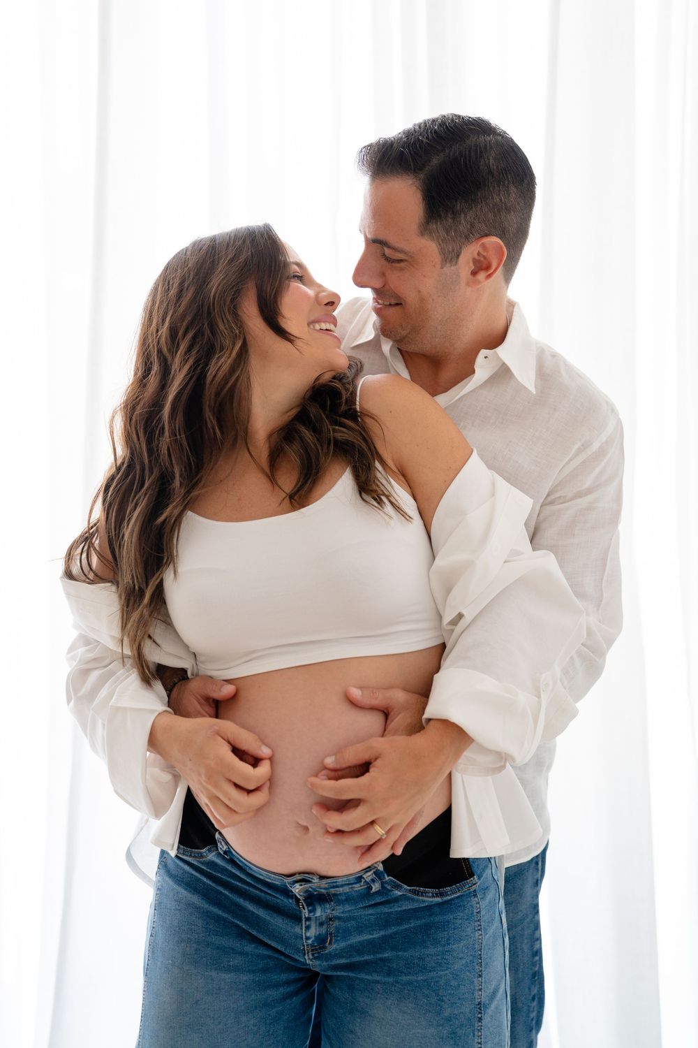 Happy couple embracing, man holding pregnant woman's baby bump, smiling at each other in front of white curtains.