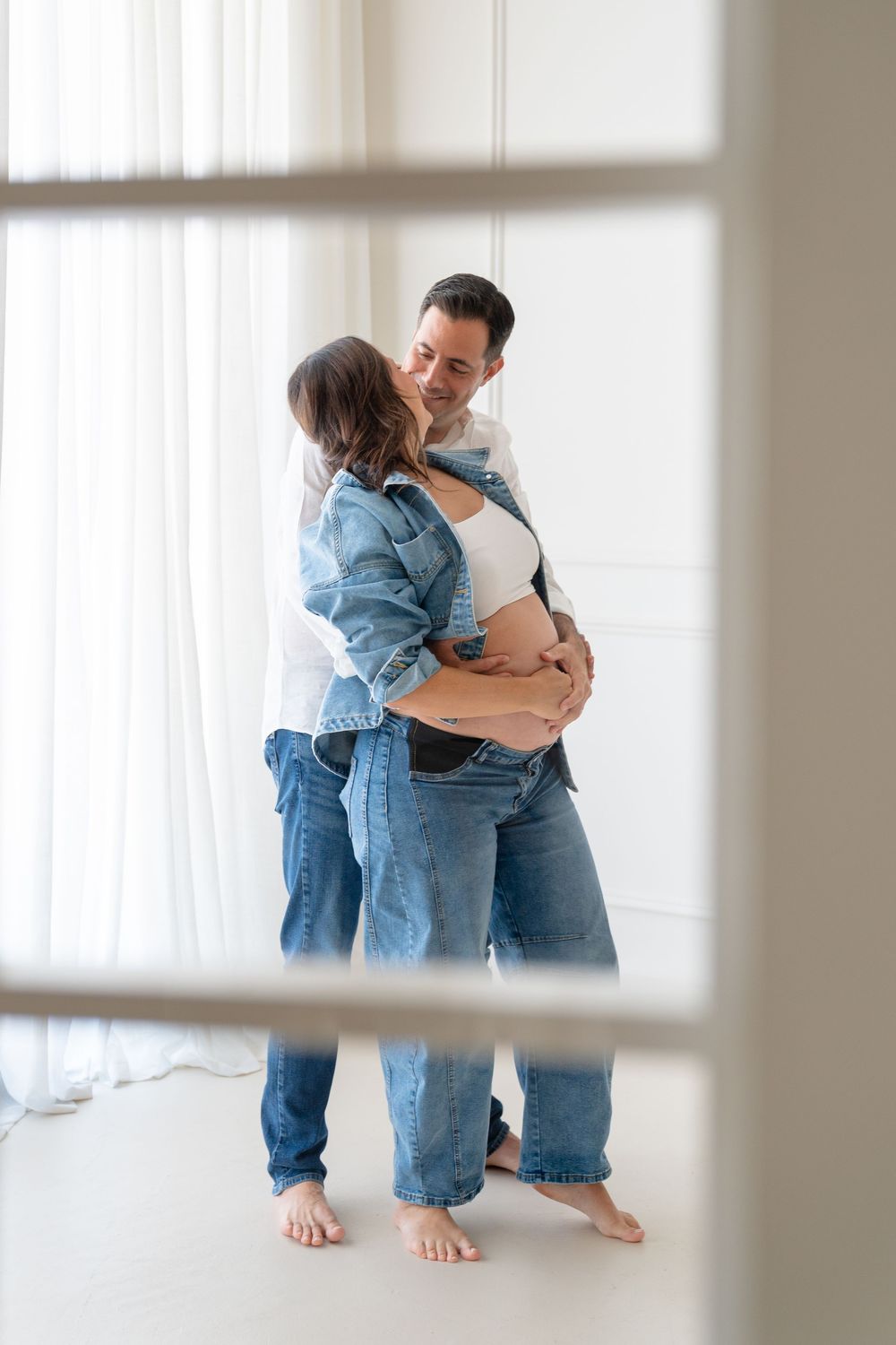 Happy couple embracing in bright white room, pregnant woman in denim jacket with partner lovingly holding her baby bump.