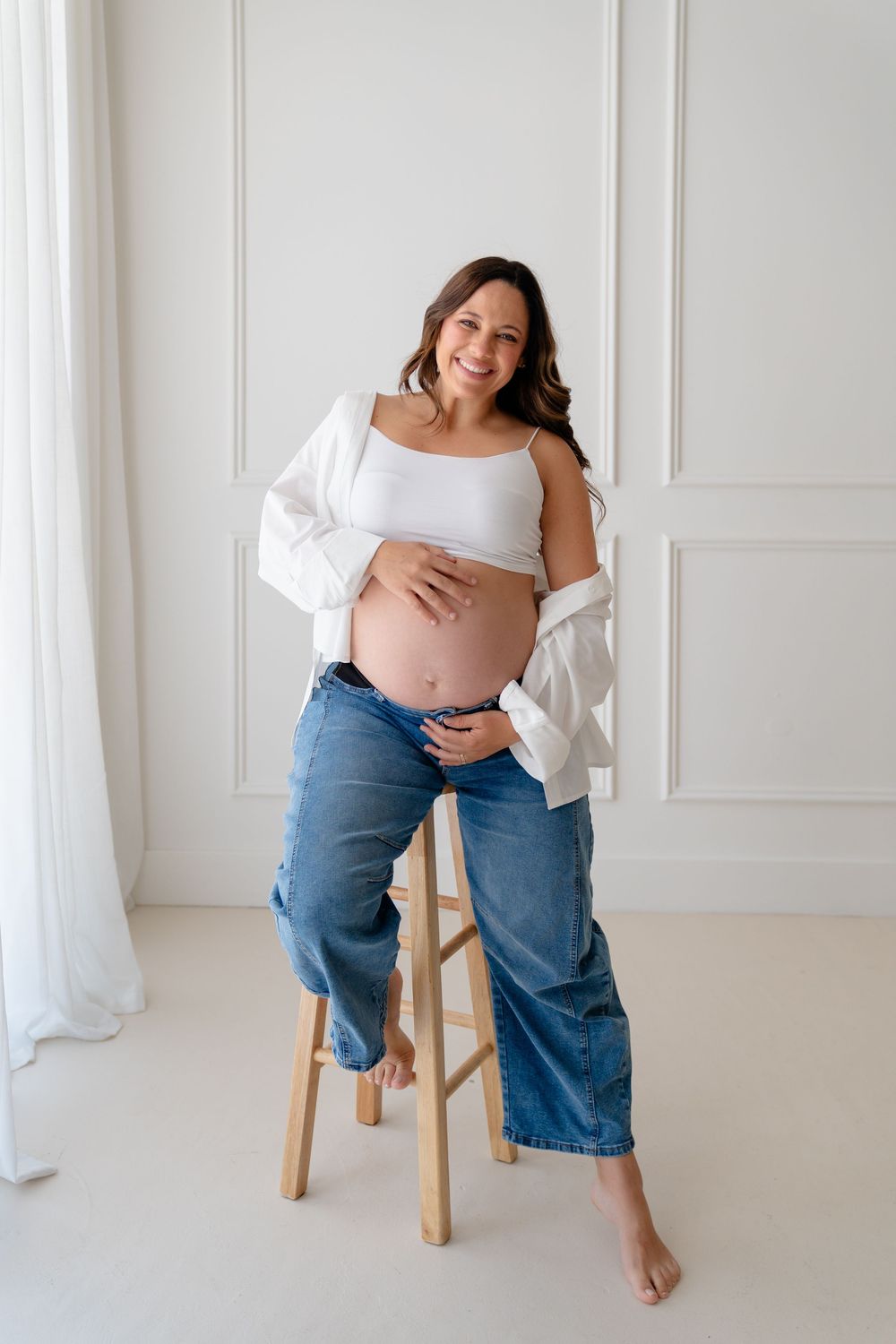Smiling pregnant woman in white crop top, open shirt, and blue jeans, cradling her baby bump while seated on a wooden stool.
