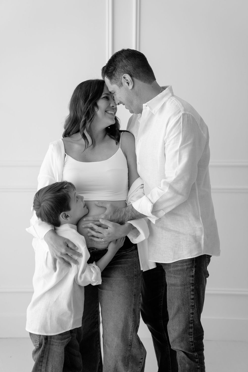 Black and white maternity photo of a couple and young boy lovingly embracing the pregnant mothers belly in a studio.