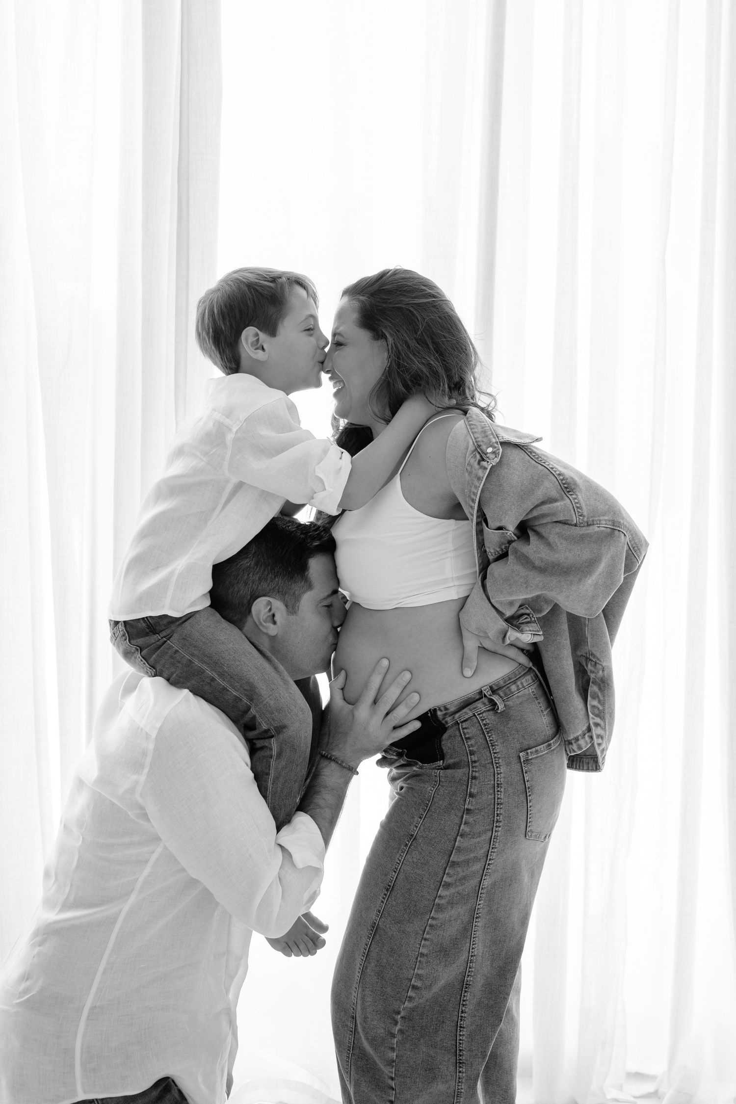 Black and white family maternity photo with pregnant woman, partner kissing belly, and young son sharing a kiss.