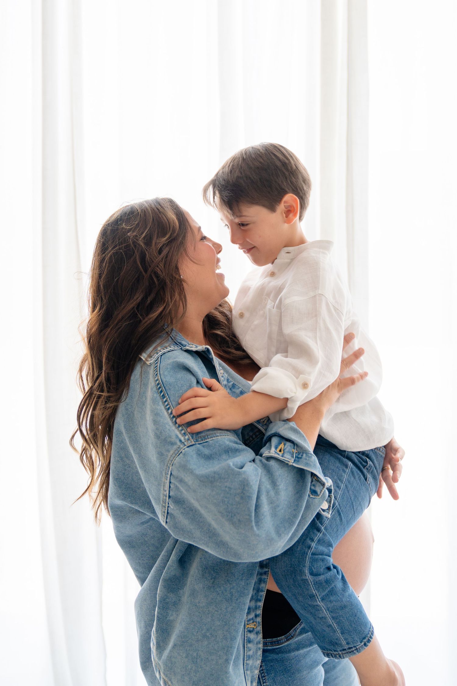 Pregnant woman in denim jacket lovingly holds young boy near white curtains.