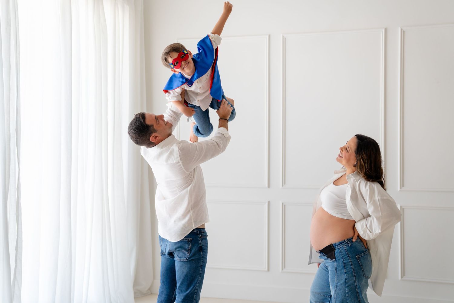 Pregnant woman watches husband lift toddler in superhero costume overhead in bright white room.