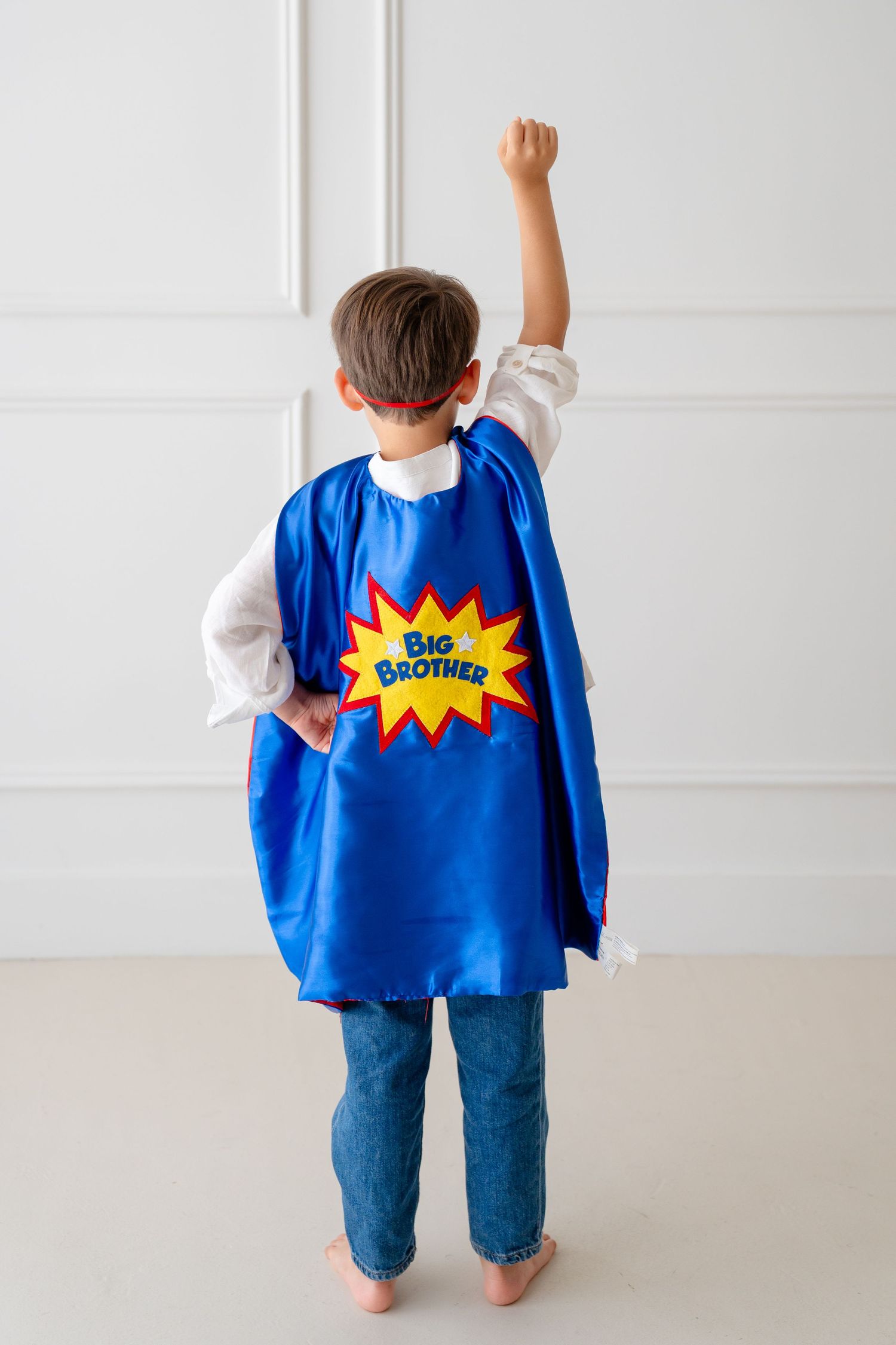 Boy in blue superhero cape with Big Brother emblem raises fist, viewed from behind.