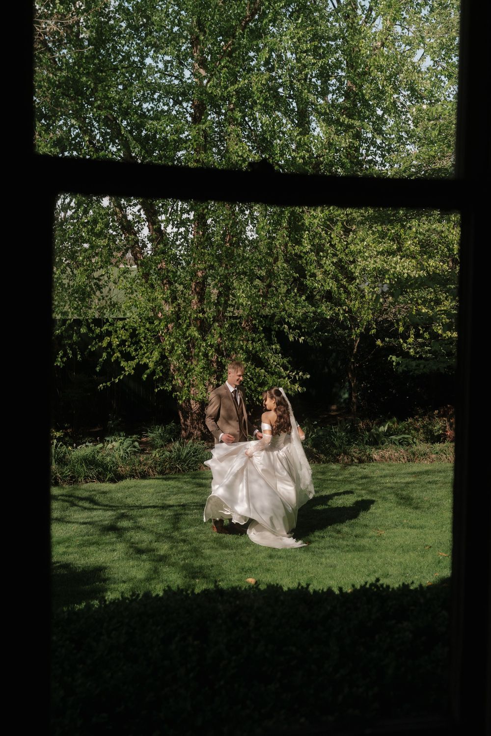Bride and groom walking hand in hand on lush green lawn, viewed through a window frame, surrounded by trees.