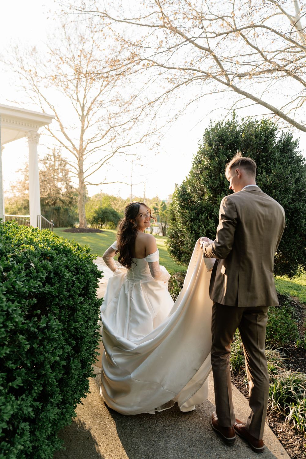 Bride in white gown smiles as groom in tan suit carries her train along a garden path at golden hour.