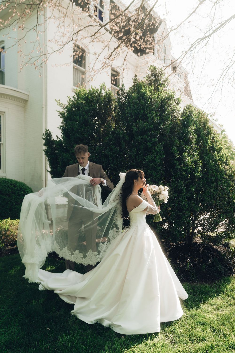 Bride in white ballgown with flowing veil and groom in brown suit pose outdoors near green hedges and white estate.