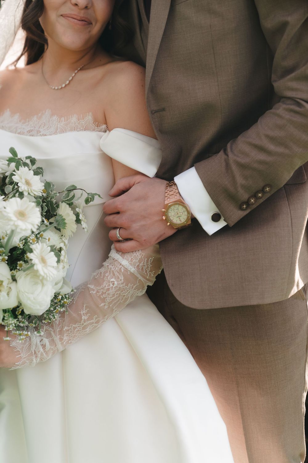 Bride in white lace gown holding floral bouquet, groom in tan suit with gold watch, wedding couple close-up.