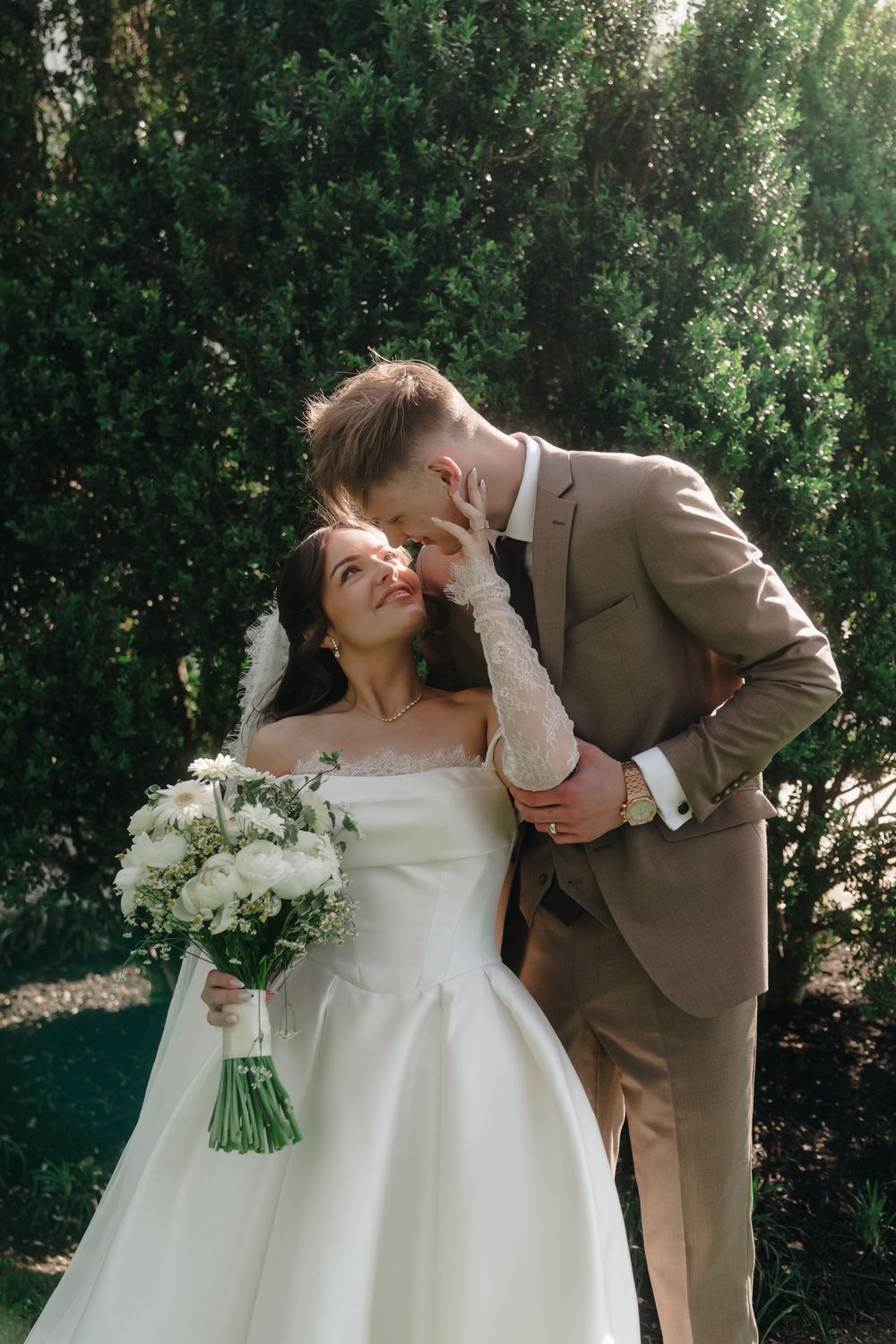 Bride in white gown holding bouquet gazes at groom in tan suit as he leans in for a kiss outdoors.