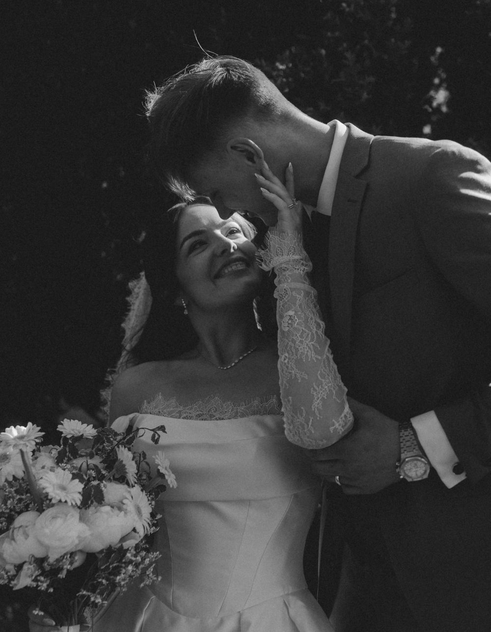 Black and white wedding photo of smiling bride in lace gloves holding bouquet as groom tenderly kisses her forehead.