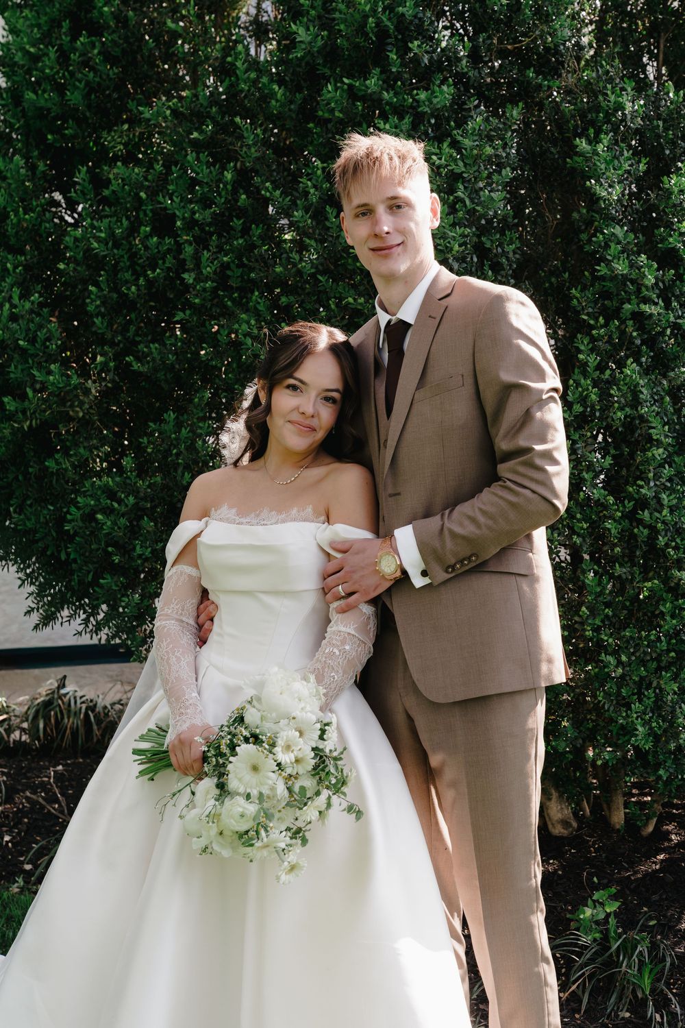 A smiling bride in a white off-shoulder gown holds a bouquet beside a groom in a tan suit outdoors.