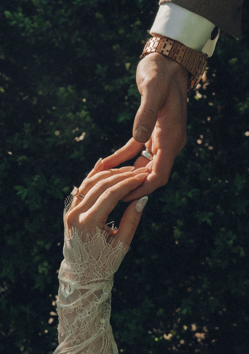Bride and groom holding hands, showing wedding rings, lace sleeve and gold watch, with green foliage background.