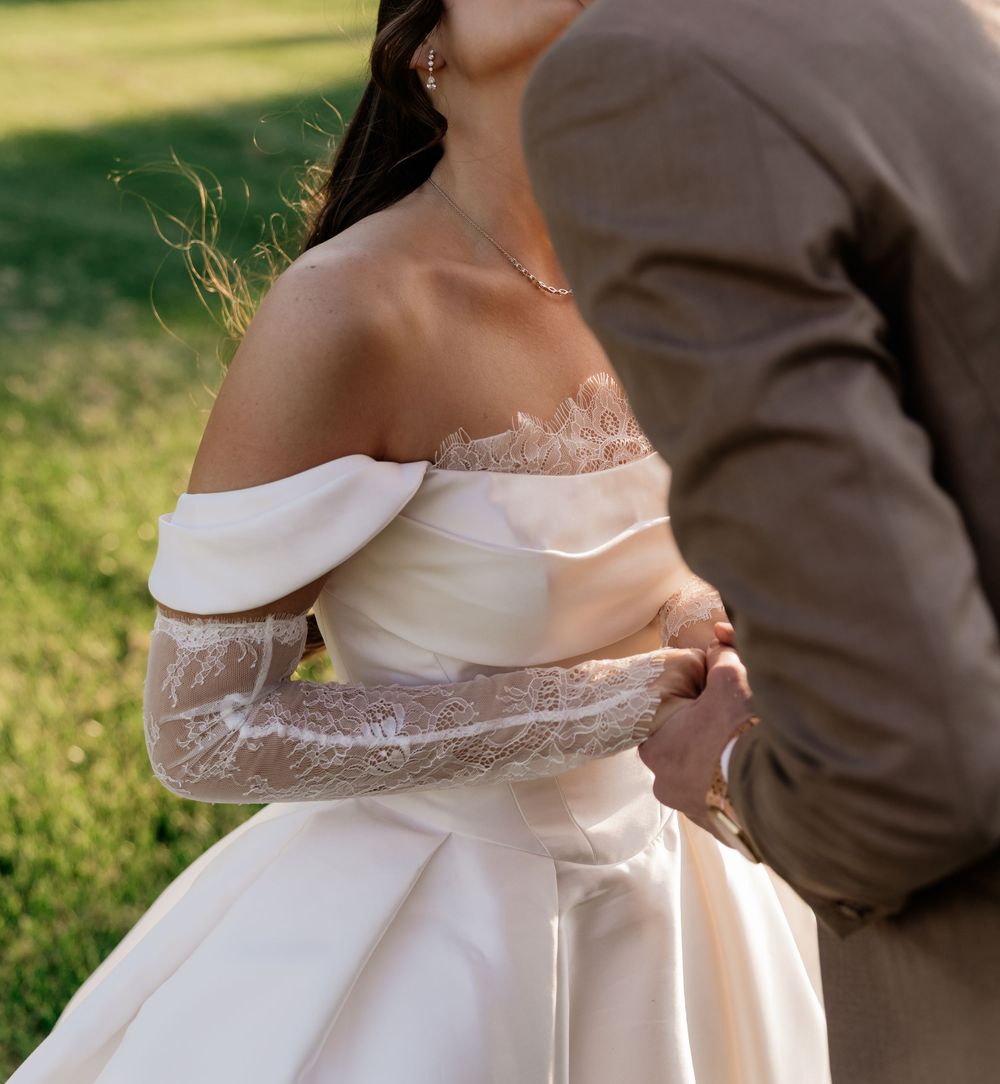 Bride in off-shoulder white gown with lace gloves held by groom in tan suit outdoors.
