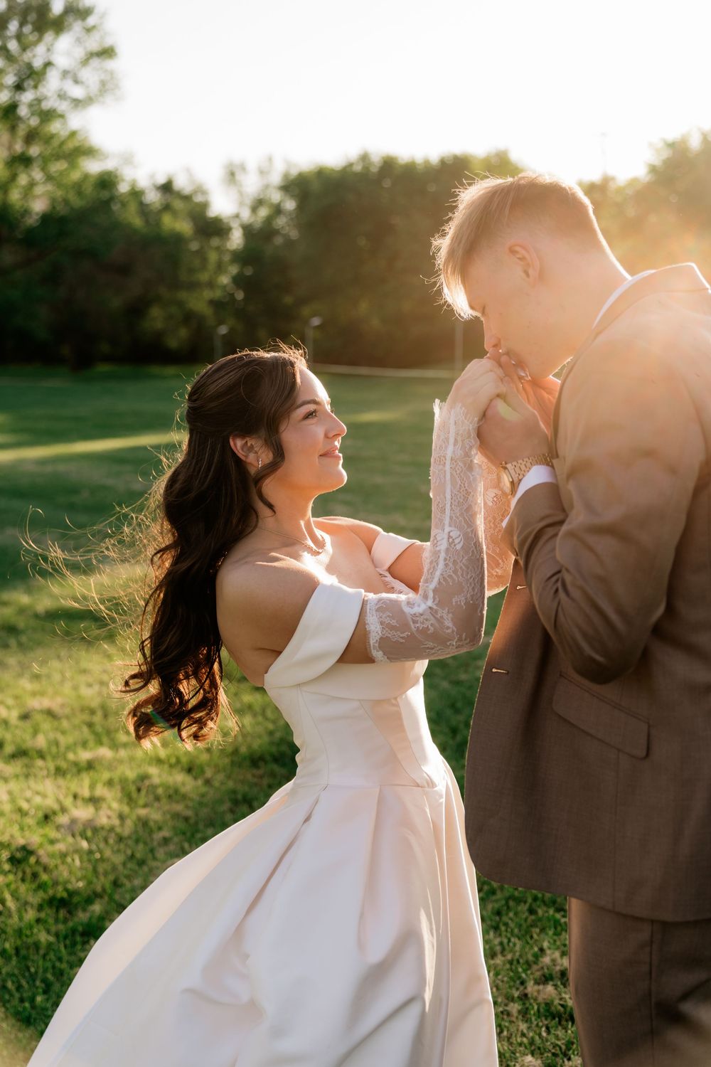 Bride in white off-shoulder gown smiles as groom kisses her hand during golden hour outdoor wedding.
