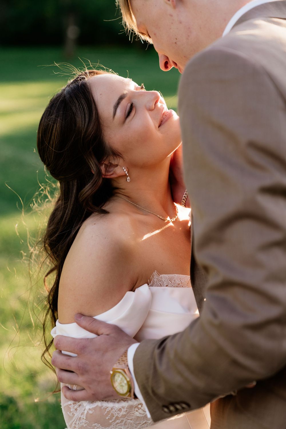 Bride gazes up at groom in golden sunlight, wearing strapless lace wedding dress outdoors.