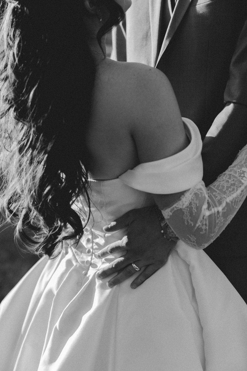Black and white photo of bride in off-shoulder gown embraced by groom with tattooed sleeve at wedding.