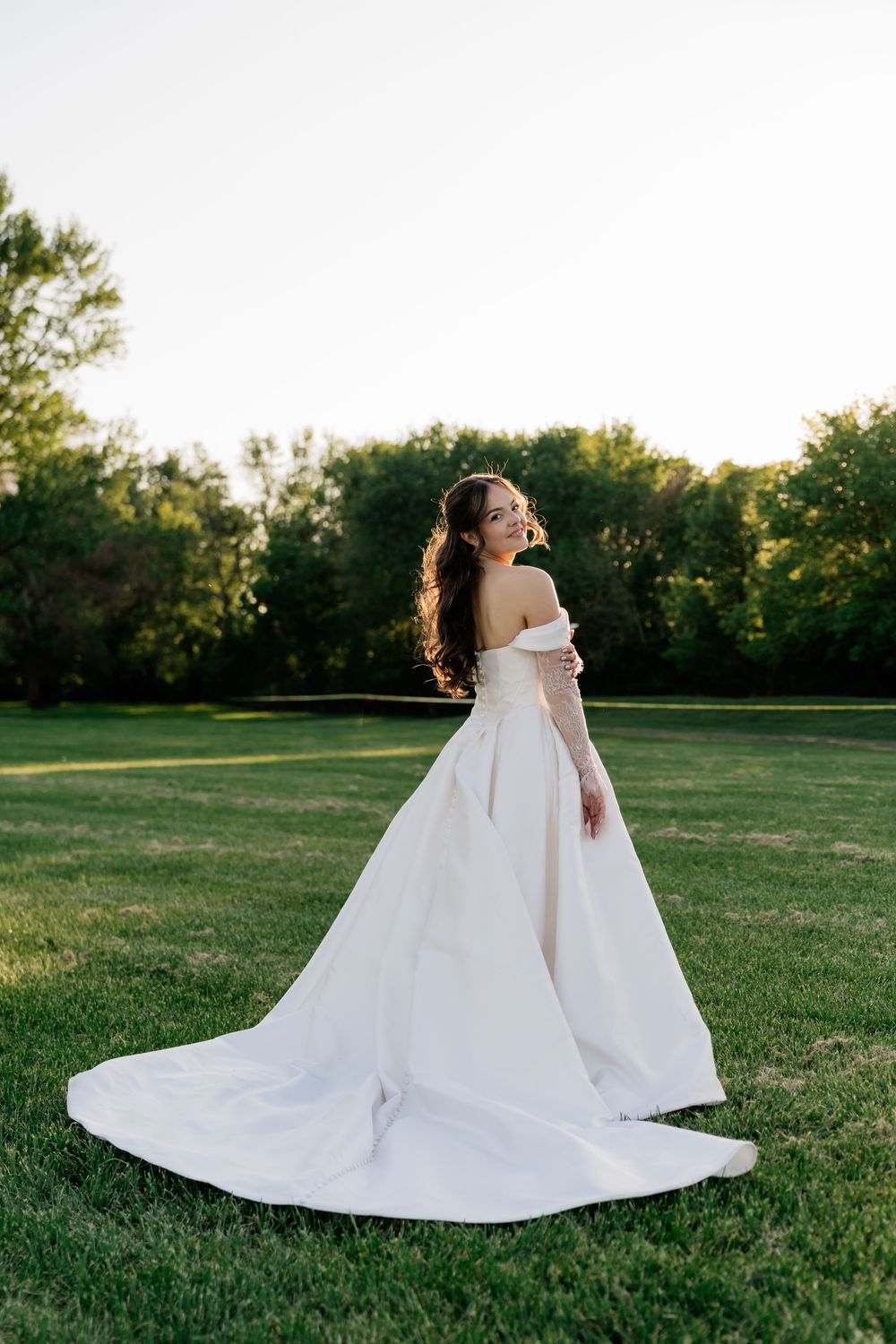 Bride in elegant off-shoulder white ball gown with long train standing on green grass surrounded by trees.