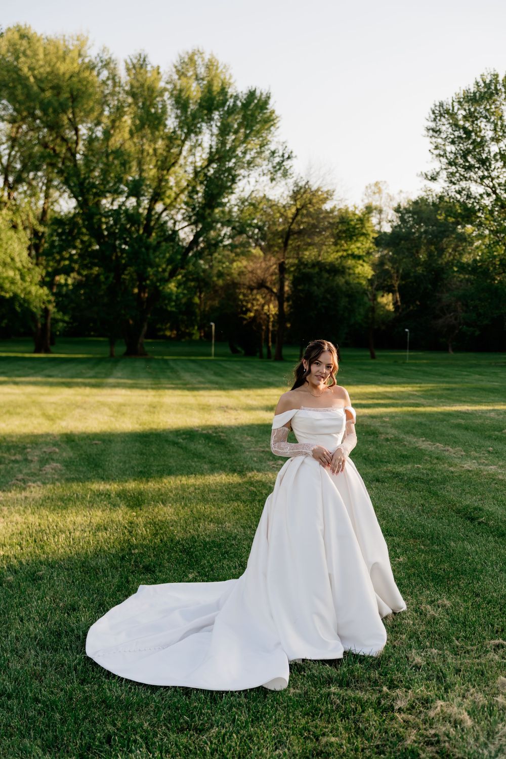 Bride in off-shoulder white ball gown with long train stands on lush green lawn surrounded by trees at golden hour.