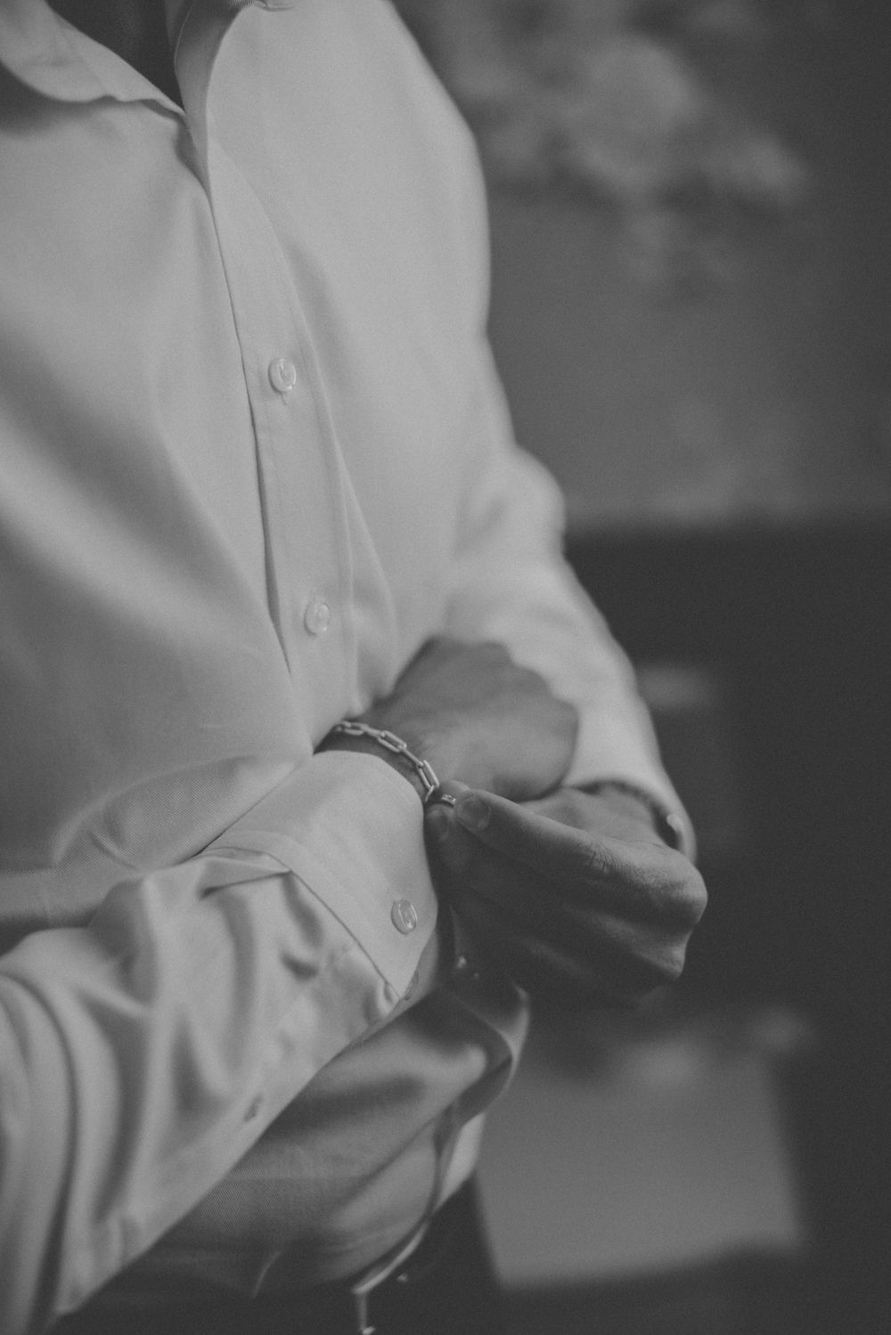 Black and white photo of a groom in a white dress shirt fastening his cufflinks with a bracelet on his wrist.