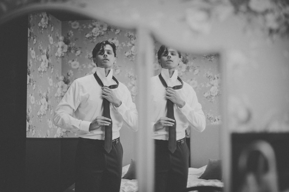 Black and white photo of a groom adjusting his tie reflected in a mirror while getting ready.