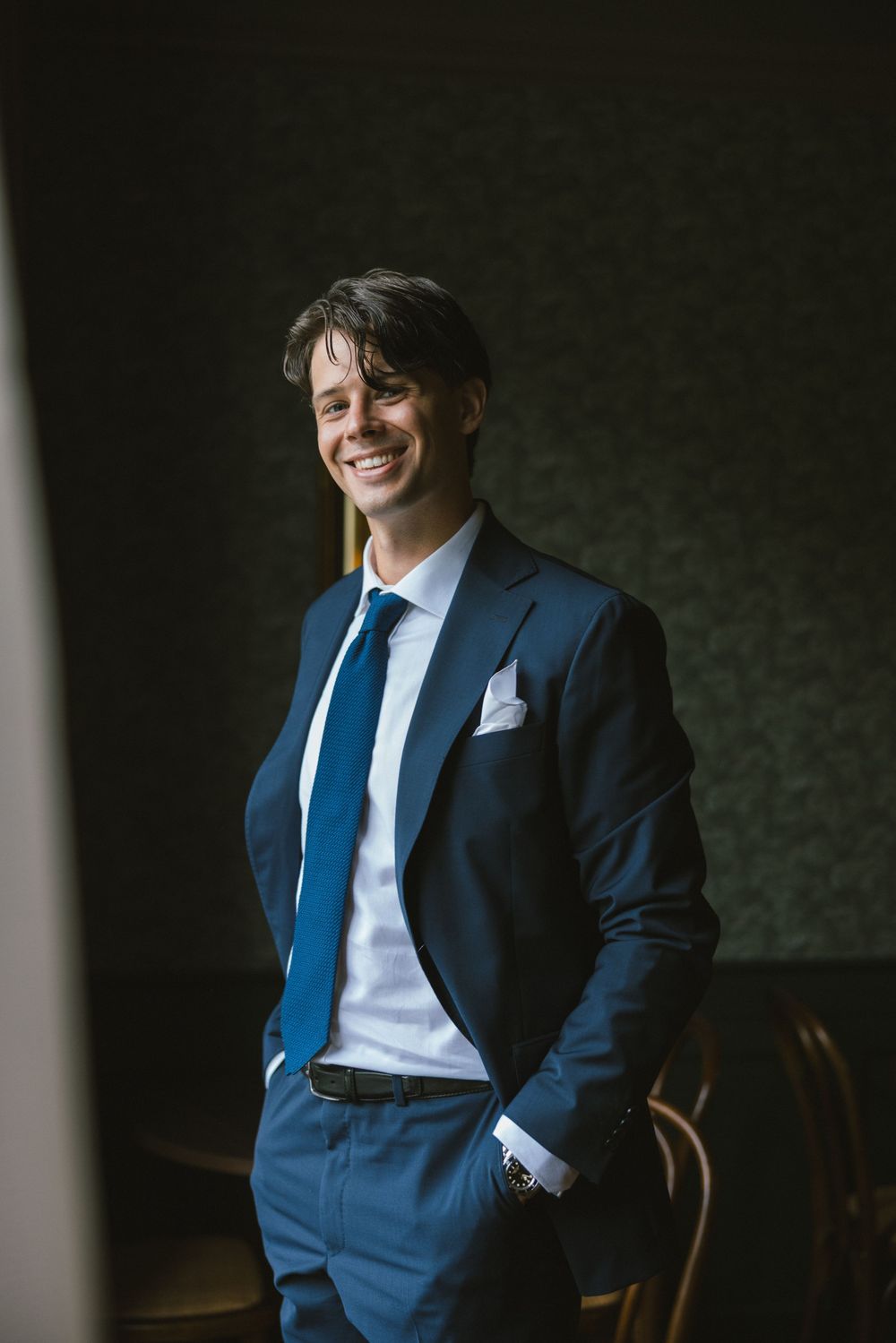 Elegant groom in navy blue suit and blue tie smiling confidently with hands in pockets.