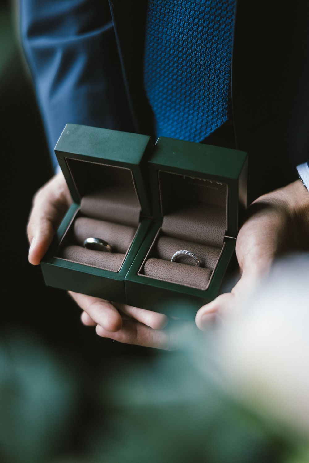Man in blue suit holding open green jewelry box with two wedding rings inside.