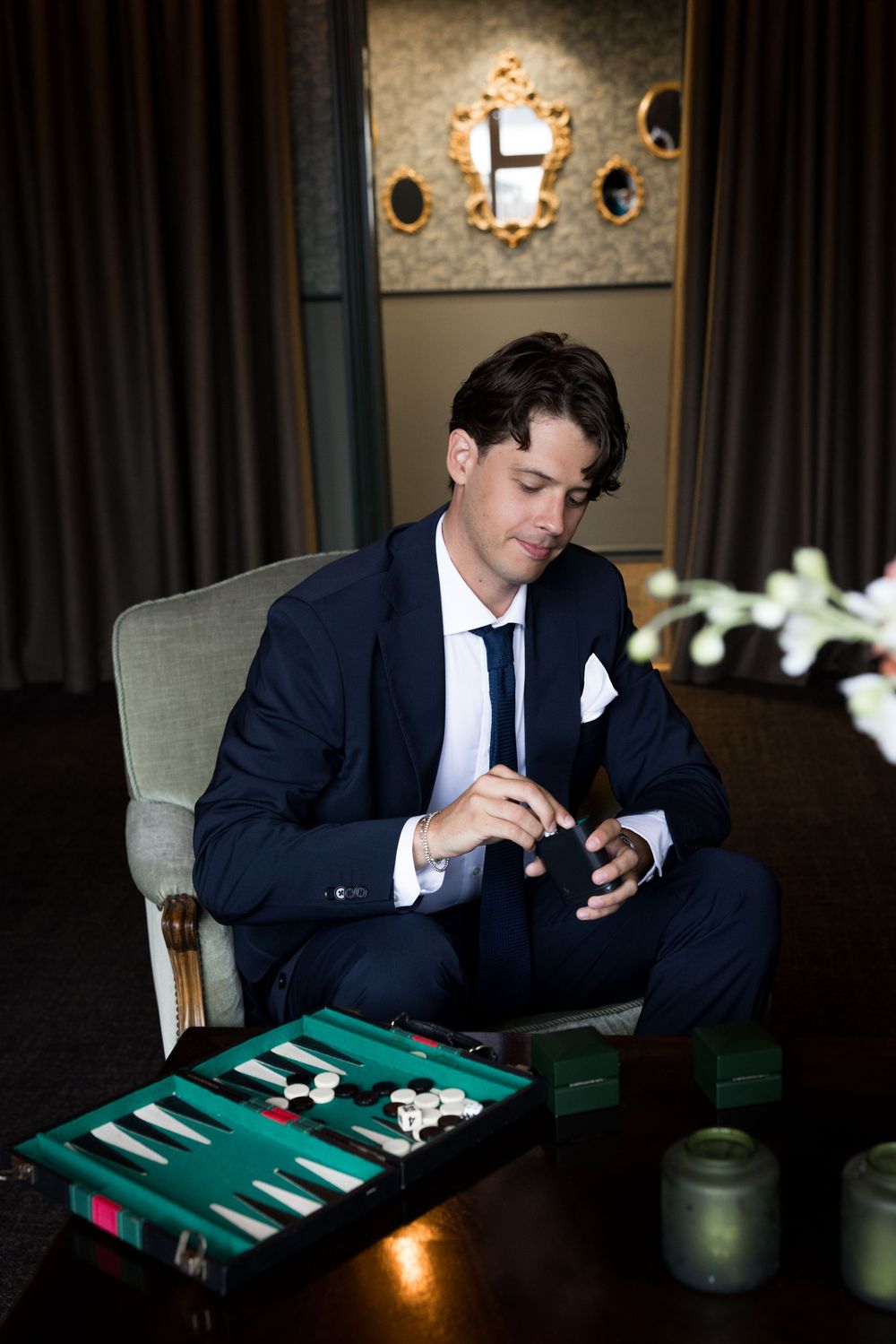 Man in navy suit playing backgammon alone in an elegant dimly lit room with ornate mirrors.