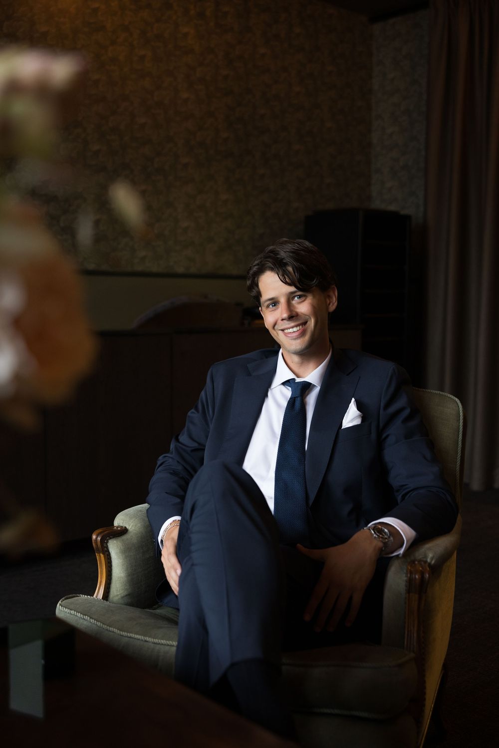 Smiling groom in navy suit sitting in vintage armchair in elegant dark room.