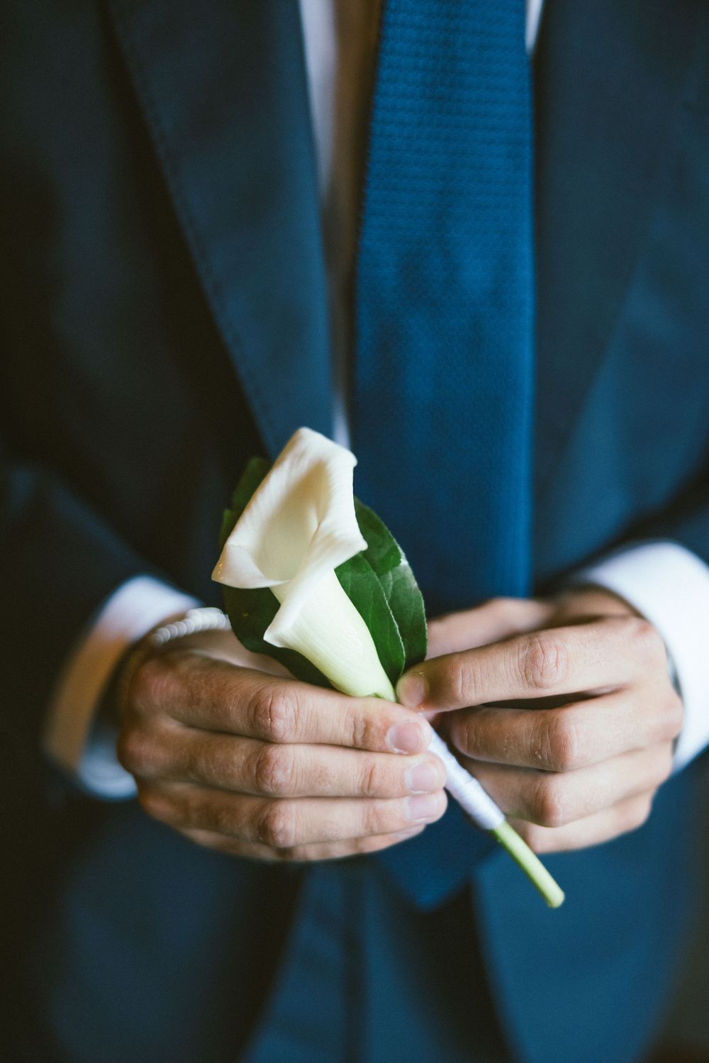 Man in blue suit holding a white calla lily boutonniere with green leaves for a wedding ceremony.