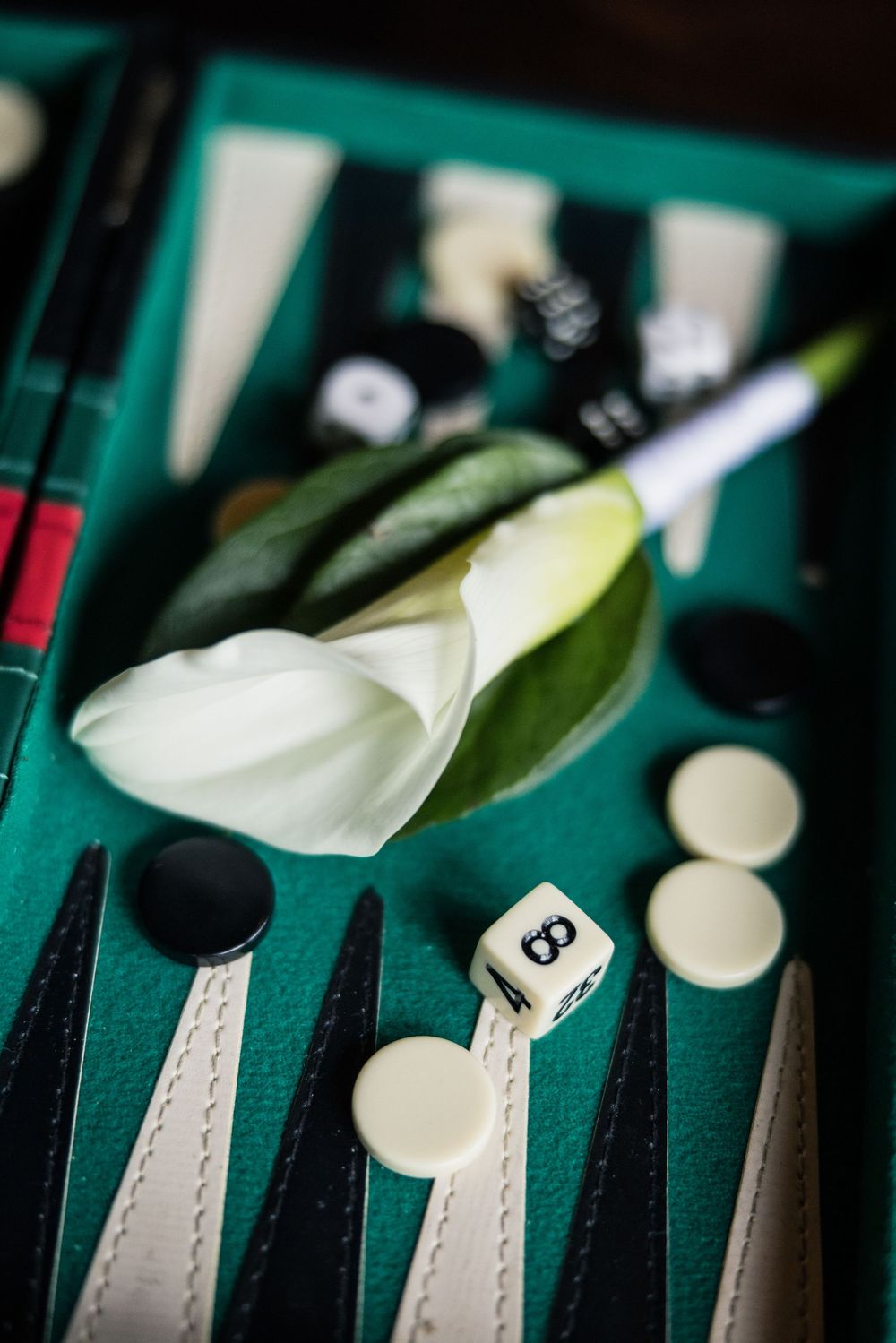 A white calla lily rests on a green backgammon board with black and white checkers and dice nearby.