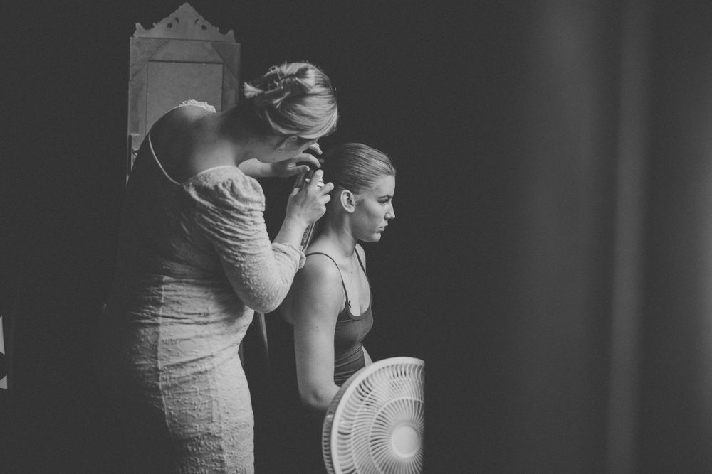 Black and white photo of a woman styling another woman's hair backstage before a wedding ceremony.