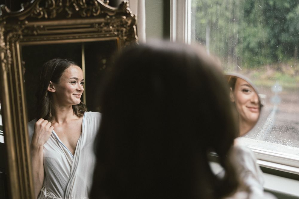 Bride smiling at her reflection in an ornate gold mirror near a rain-streaked window on her wedding day.