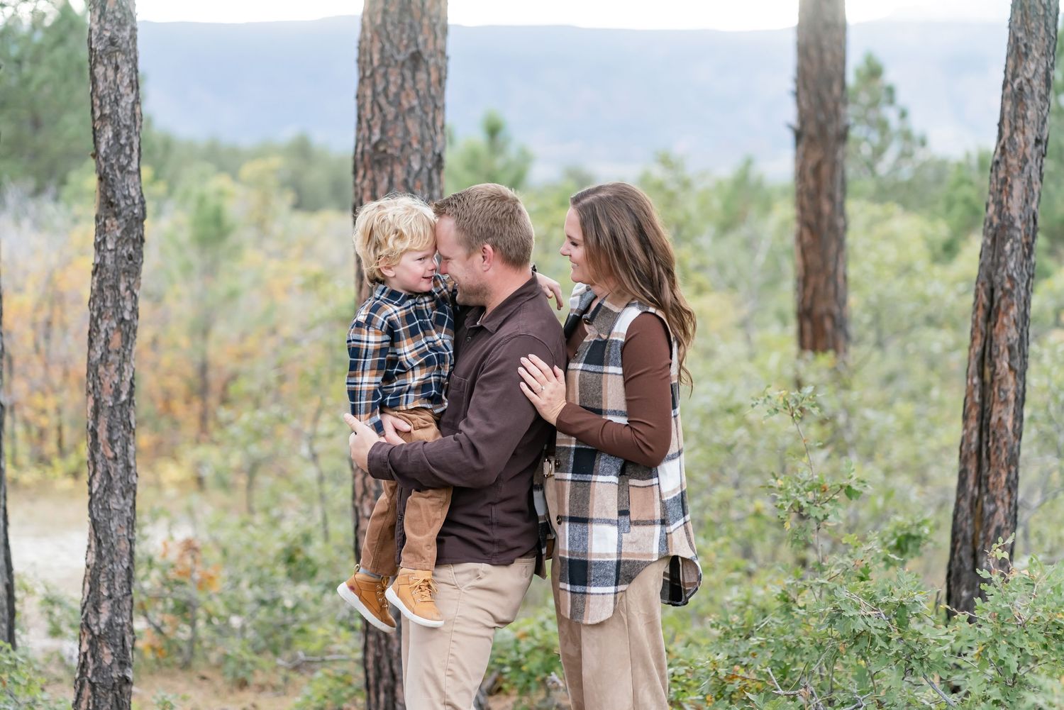 A family of three poses outdoors among pine trees, with parents holding their toddler in a warm autumn forest setting.
