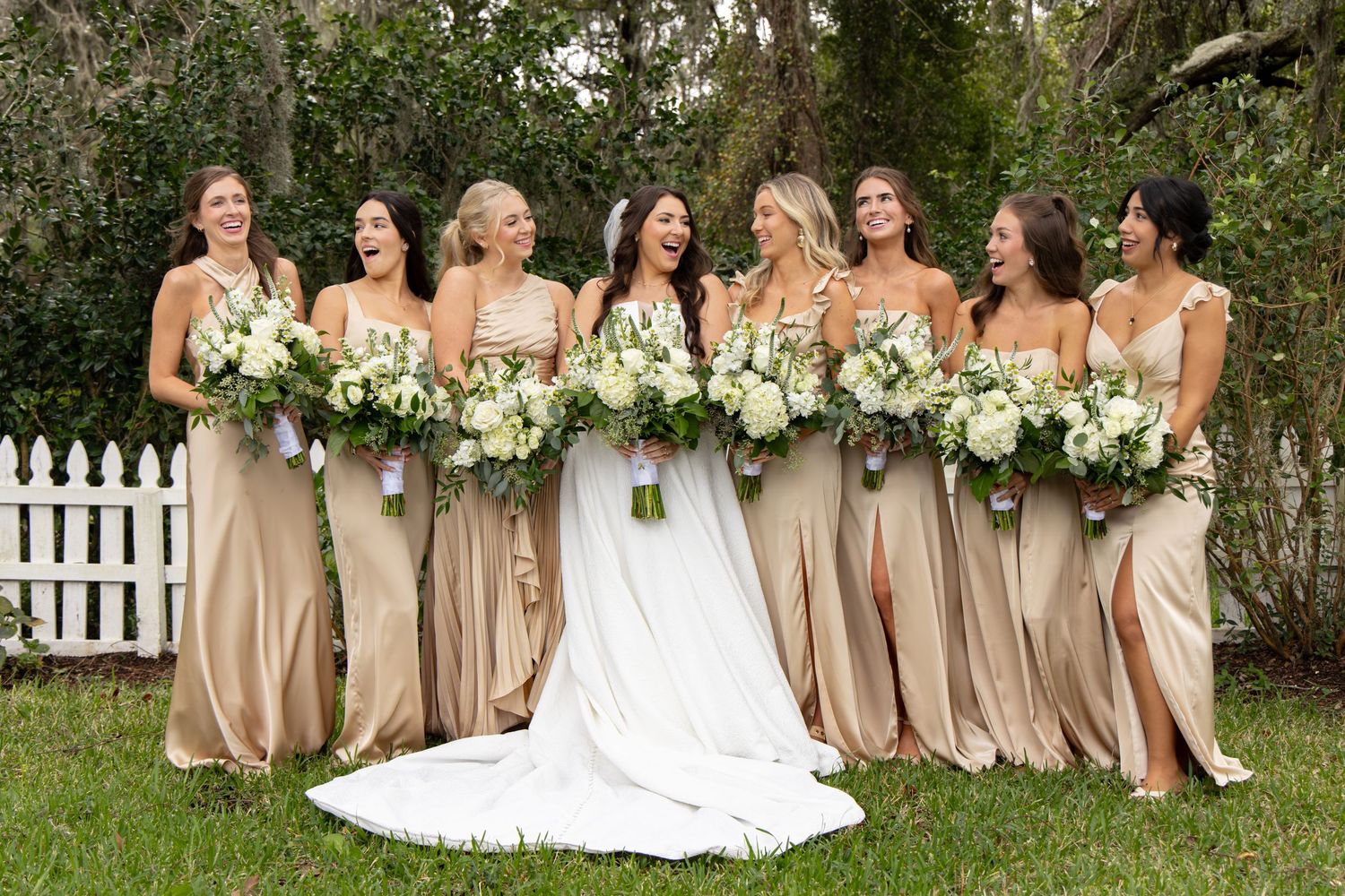 Bride in white gown with bridesmaids in champagne satin dresses holding white floral bouquets outdoors.