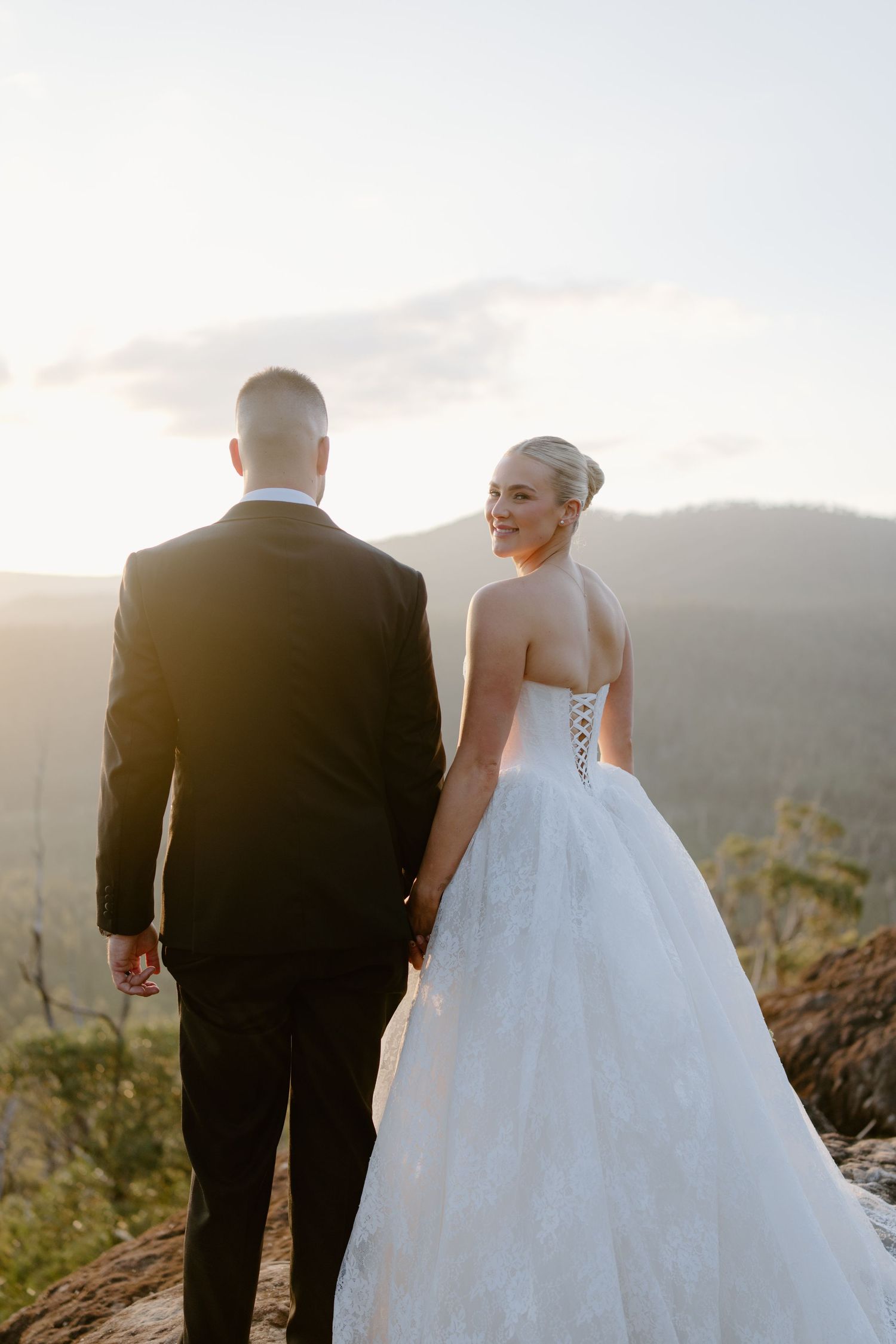 Bride in white lace ball gown and groom in black suit hold hands on mountain cliff at golden hour sunset.