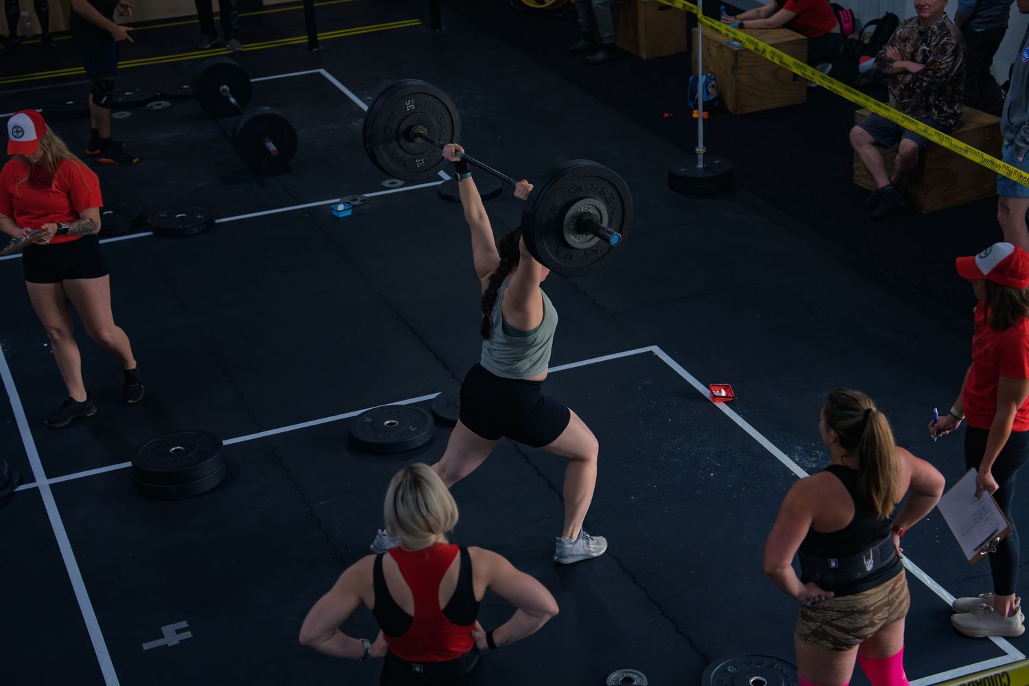 An athlete locks a loaded barbell overhead in a lunge at a gym competition, judges in red shirts watching as other lifters wait nearby