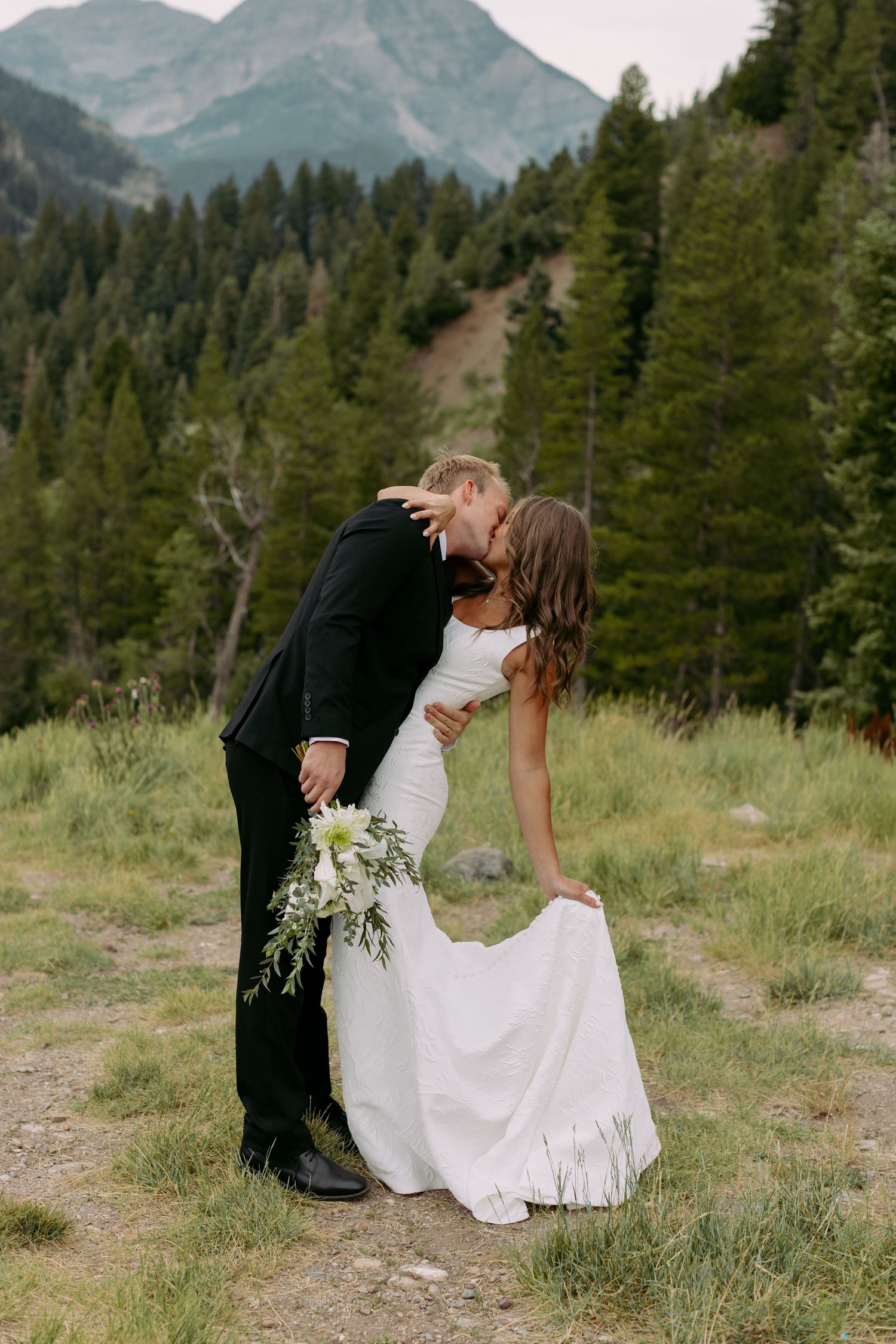 A bride and groom share a romantic kiss in a mountain meadow, surrounded by pine trees, as she holds a white bouquet.