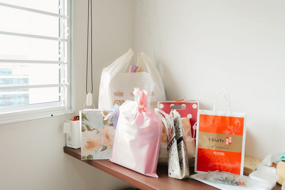 Assorted gift bags and wrapped presents arranged on a wooden shelf near a bright window.