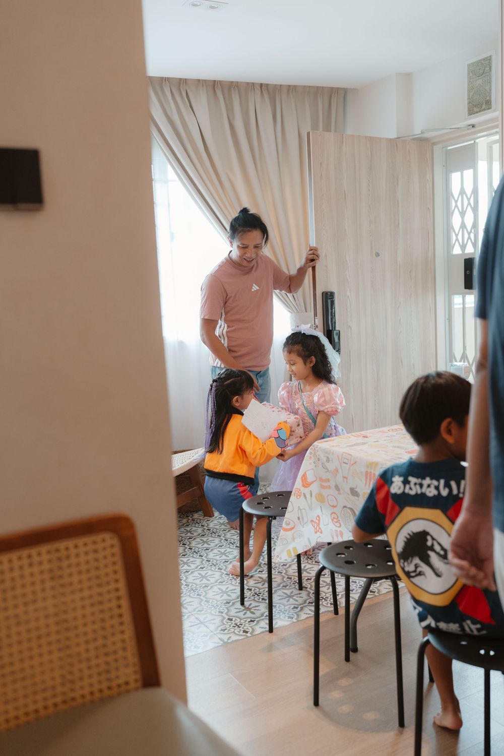 Adult and children gathered around a table near curtained window in a bright indoor setting.