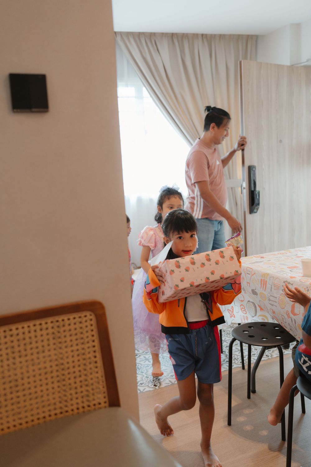 Young boy carrying a pillow walks through a room while family members follow behind in a cozy home interior.