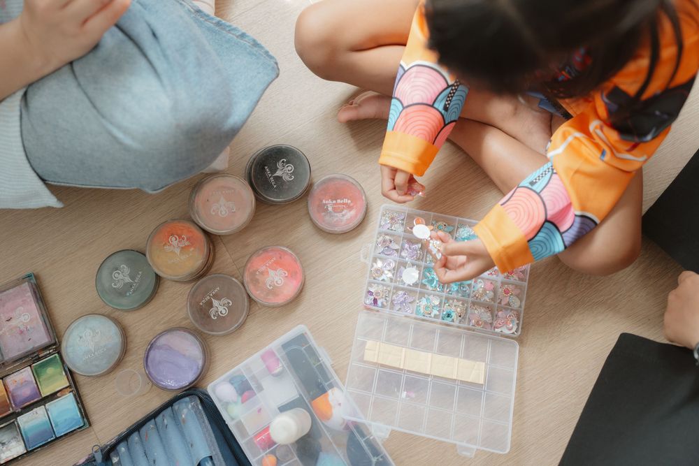 Two children sit on floor playing with colorful makeup palettes and craft supplies in a plastic organizer box.