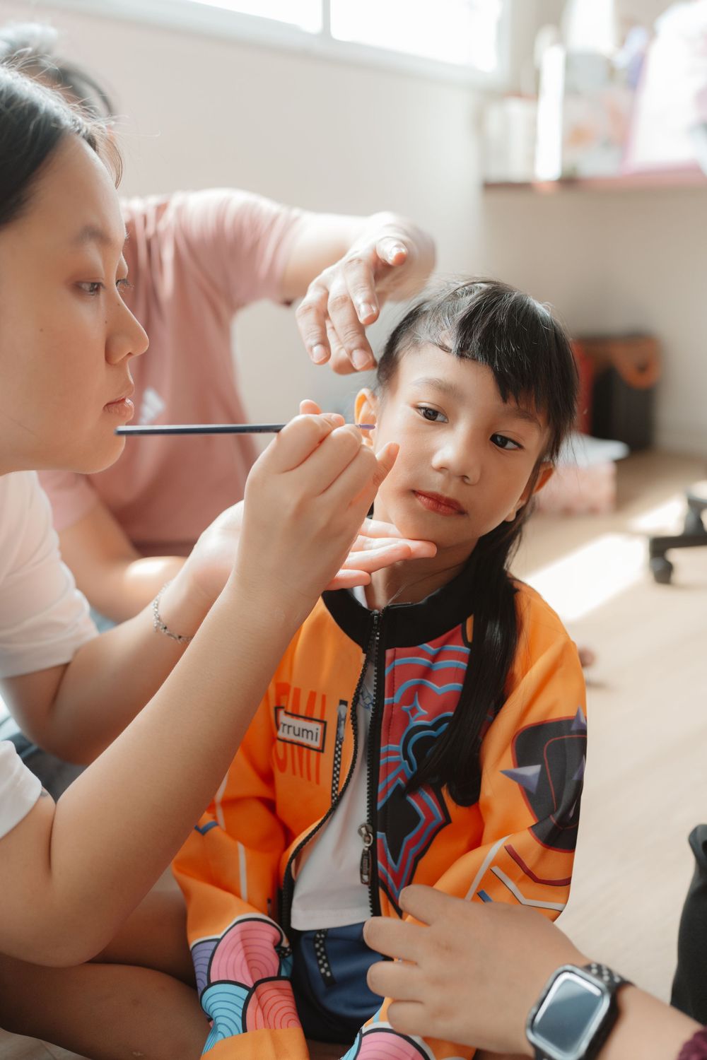 Woman applying face paint to a young girl in an orange jacket during a kids party.
